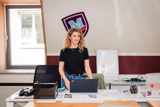 Een medewerker staat achter een bureau met een laptop, onder het schild van Miles