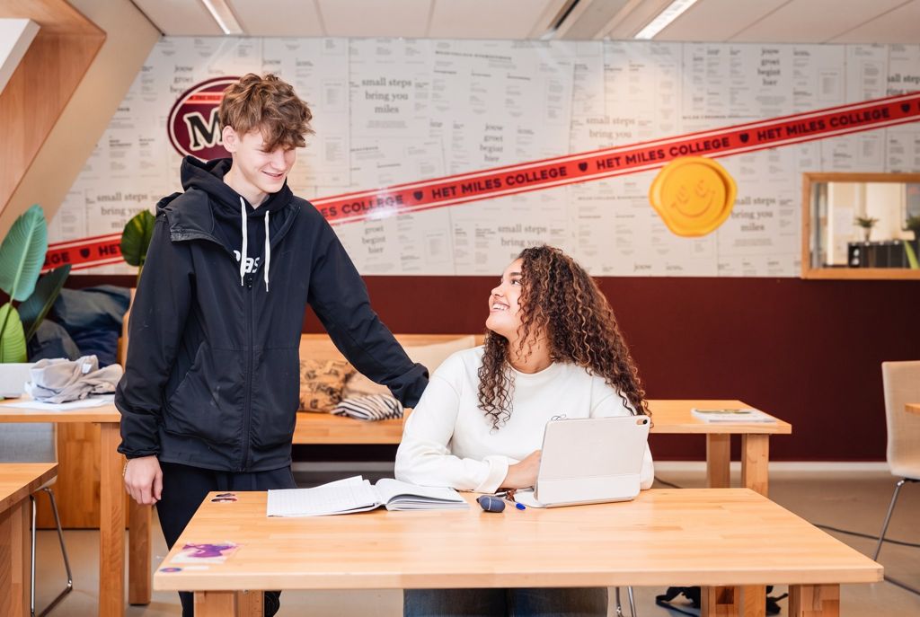 Een mentor buigt zich staand over de tafel om een leerling te helpen met haar werk