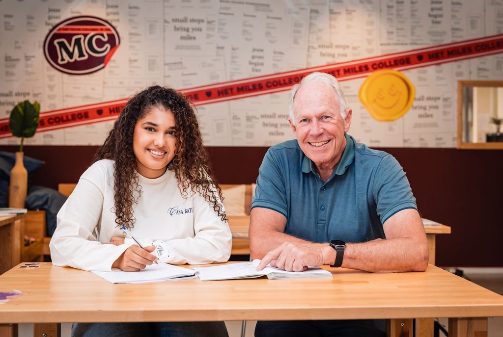 Een leerling en haar mentor lachen samen aan tafel, met het Miles-logo op de muur