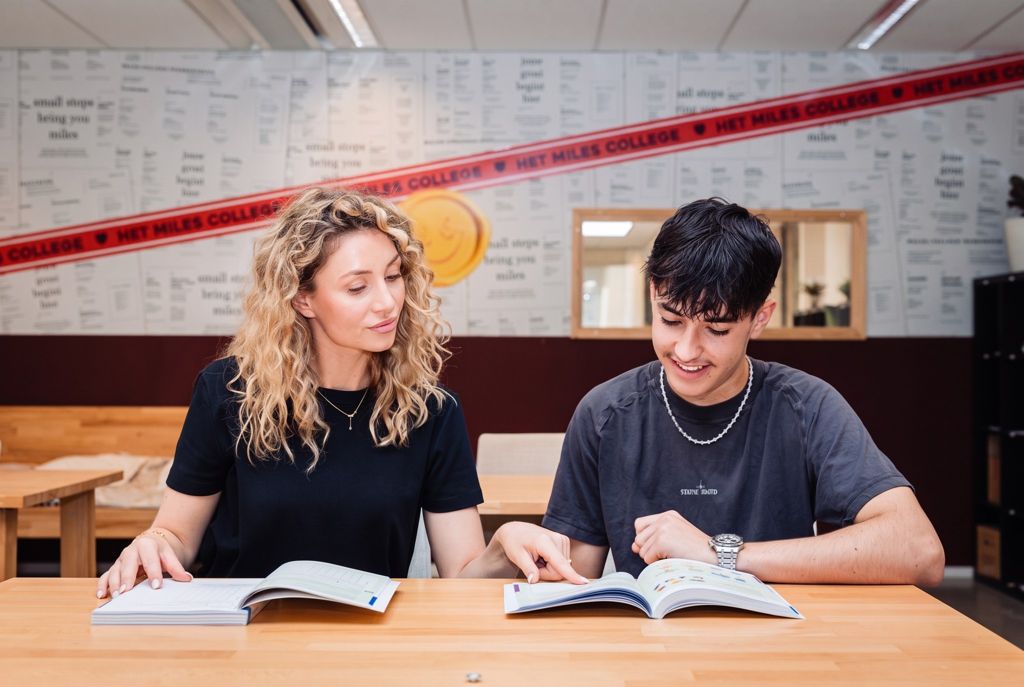 Een docente en een leerling zitten met boeken aan tafel en bespreken de stof